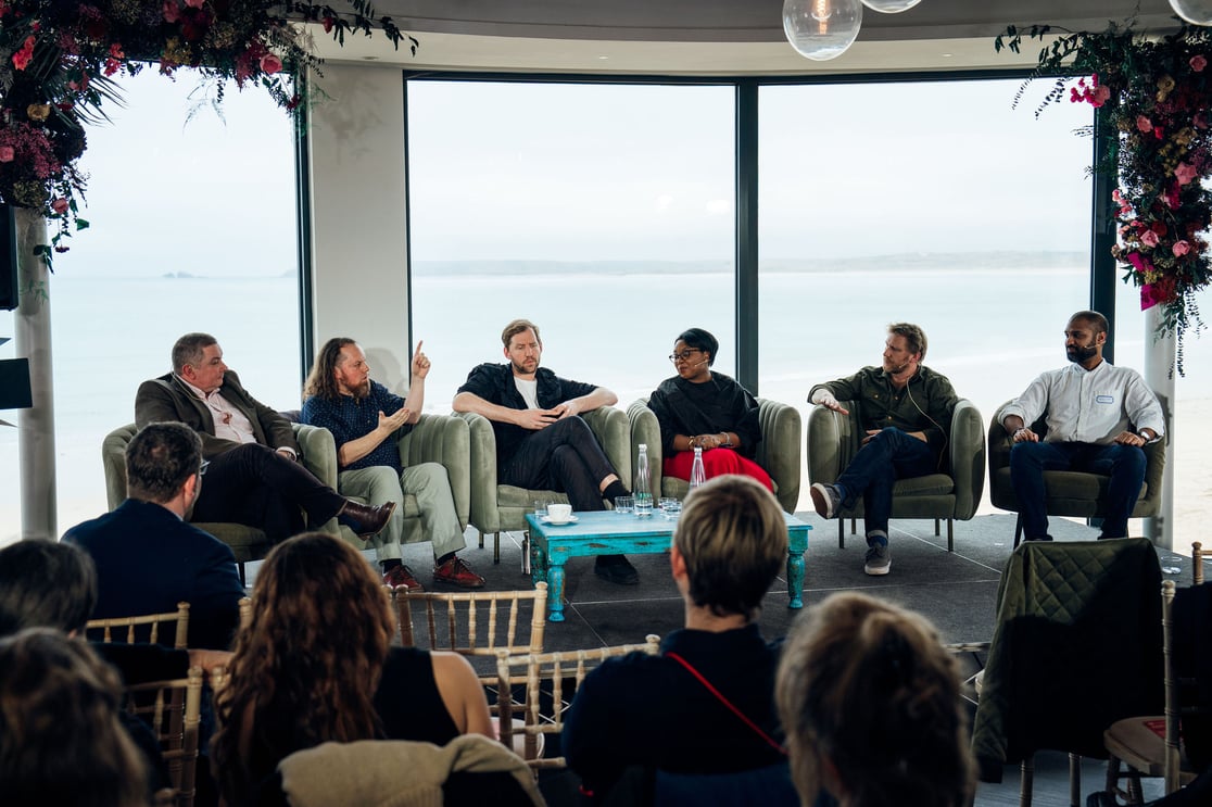 Six people sit in armchairs on a stage, with a backdrop of the ocean.