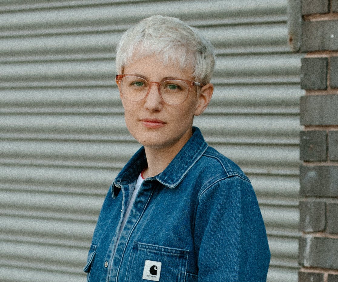 Loran has short bleached blonde hair and wears glasses and a denim jacket. She stands against a pale, shutter background.