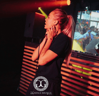 Kimmy wears a black t-shirt and has long blonde hair tied in a ponytail. They hold a microphone to their throat and stand on a stage under orange lights.