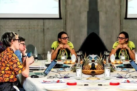 Mirrored image. People sit around a big table cluttered with laptops, papers and water bottles. Each person wears glasses. One person wears a bright green top, another wears a pale blue top and the last wears an orange patterned shirt. They are against a grey concrete background and windows.