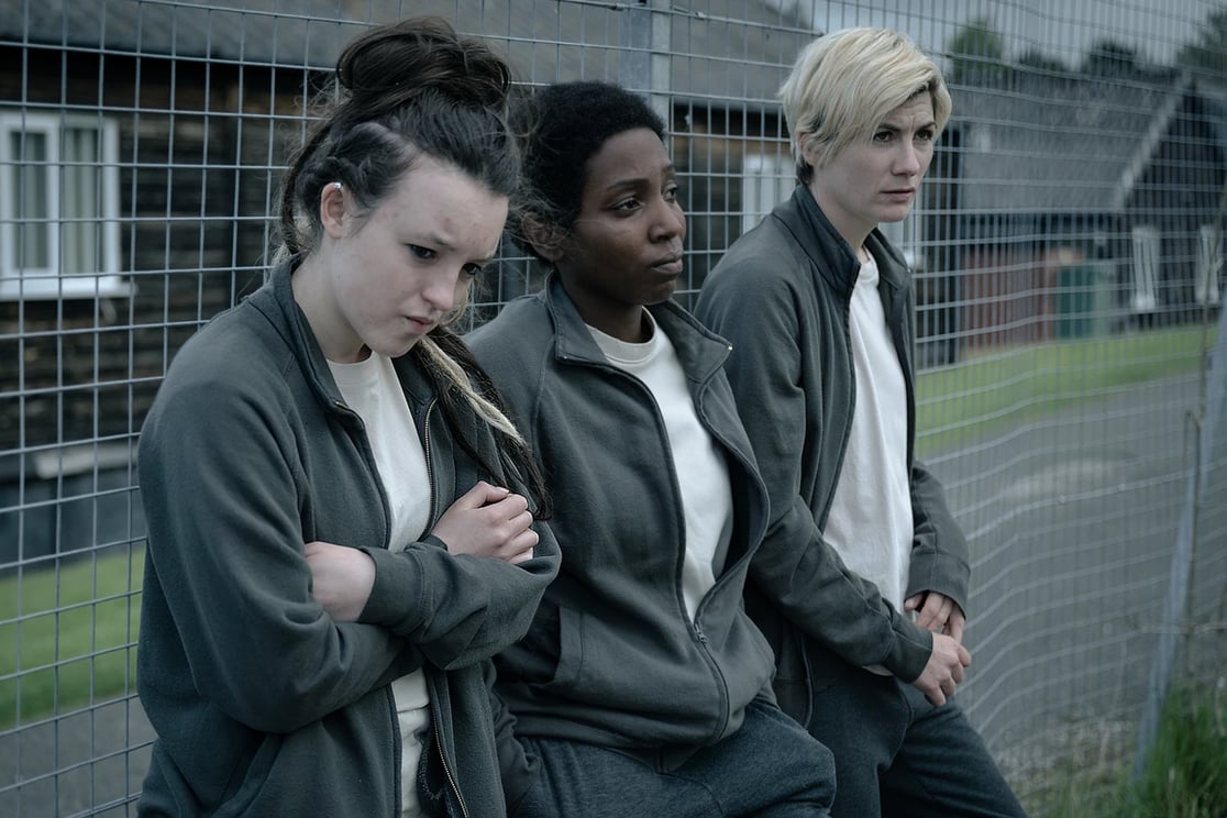 Three people leaning against a chain fence. They are all wearing grey joggers, beige t-shirts and dark green fleeces. The first person on the left has dark brown hair tied up in a messy bun, the second person has a short black afro and the third person has a blonde pixie cut.