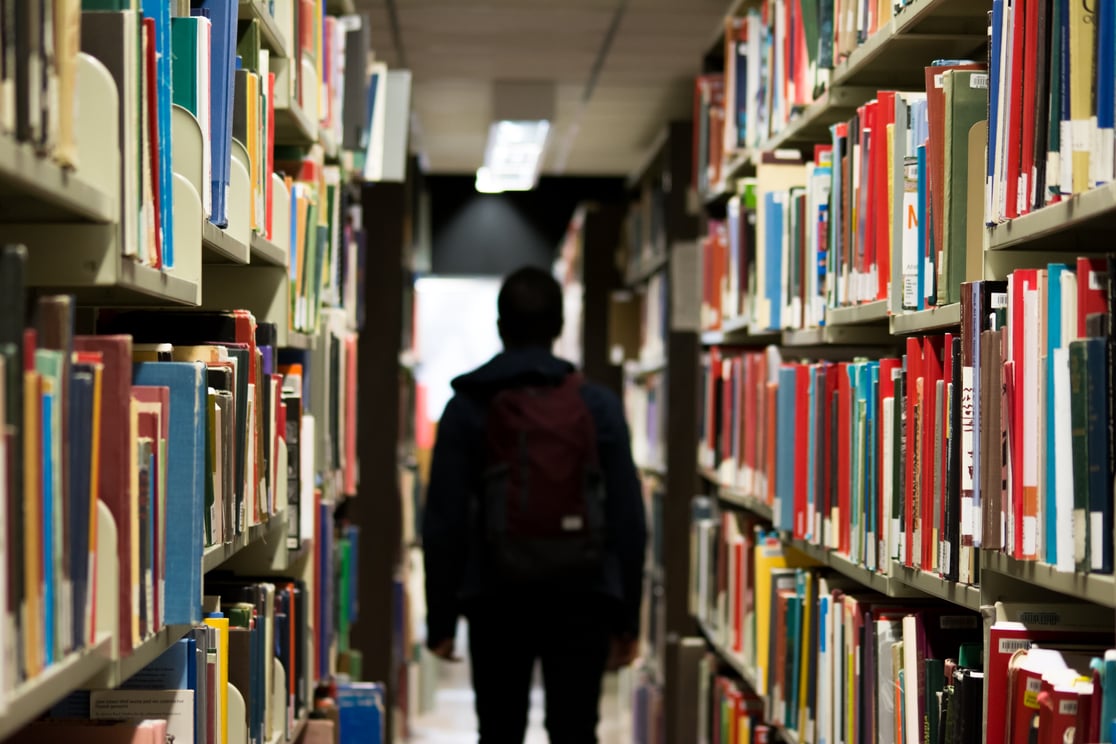 Person with a backpack walking between two rows of bookshelves.